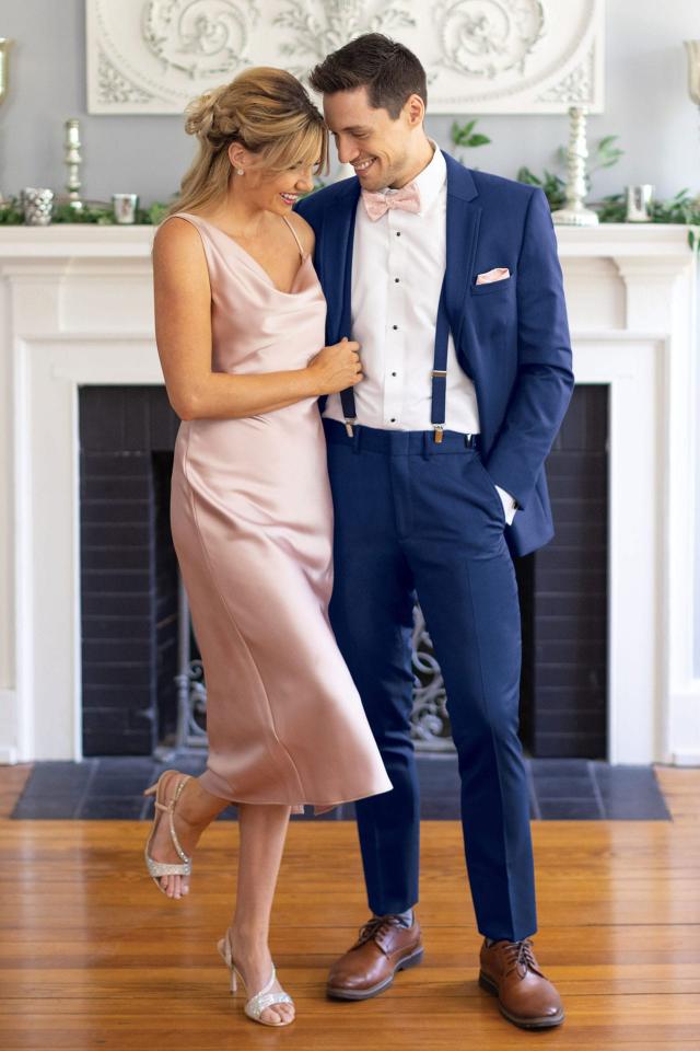 man wearing Michael Kors Blue Performance Suit and matching colored suspenders standing next to woman in blush colored slip dress holding onto the man's suspenders