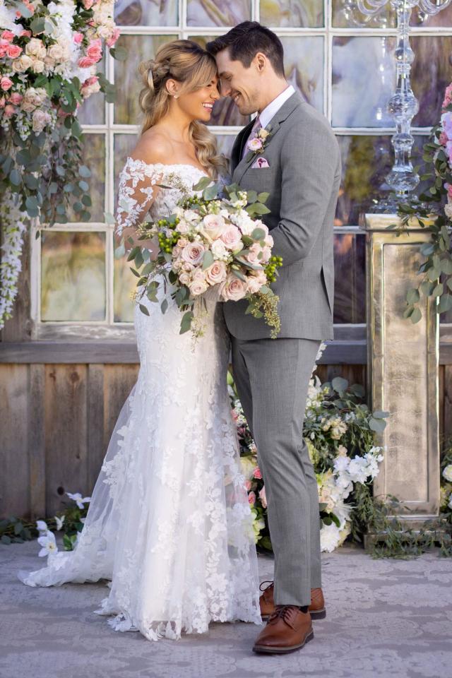 bride and groom facing each other with foreheads touching. Standing in front of stained glass windows with large displays of flowers on either side of them.